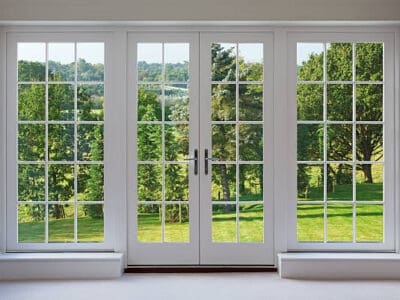a set of finely crafted wooden Georgian style windows with doors set in a white frame, with white sills and caramel coloured walls. The view through the windows are towards a beautiful  countryside garden.